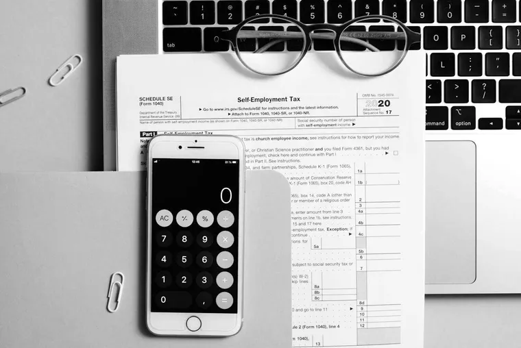 A self-employment tax form on a desk with reading glasses, a phone calculator and a laptop